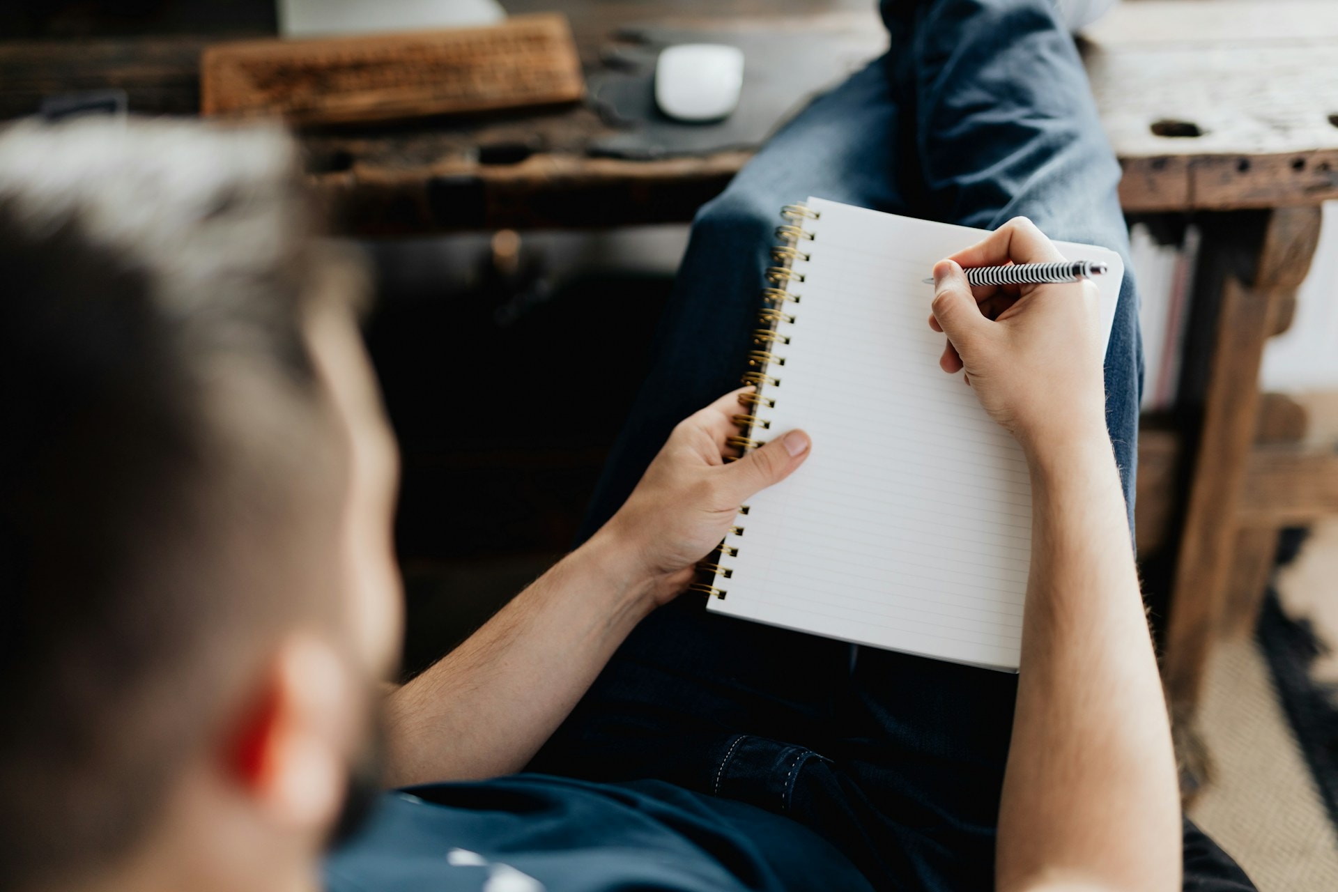 Stock image of a man lounging on a chair with his feet up on a nearby table. He has a notebook and a pen in his hands, poised to write. This is to illustrate how writers get paid to write poetry and how freelance poems make money by selling their poetry online.